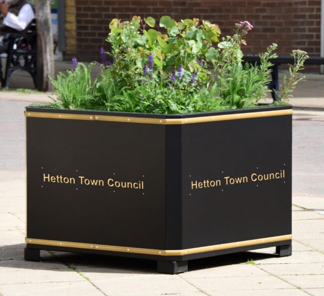 Large black planter labeled Hetton Town Council, filled with green plants and flowers.