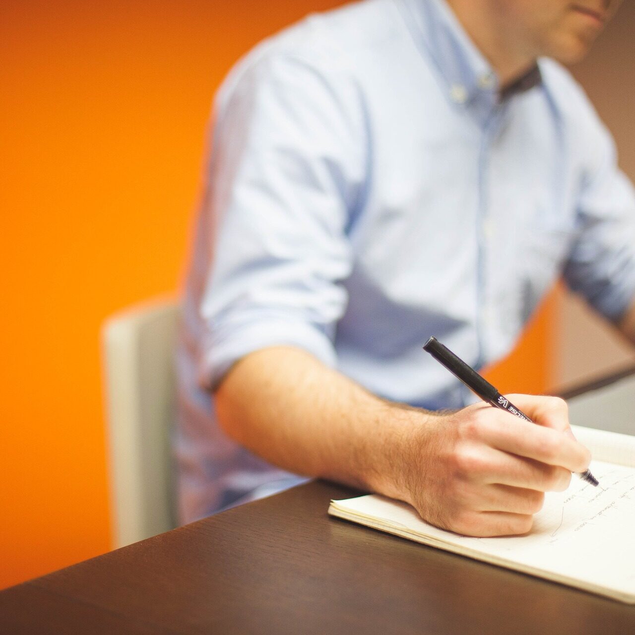 Person in a blue shirt writing in a notebook beside a laptop on a desk, orange wall behind.
