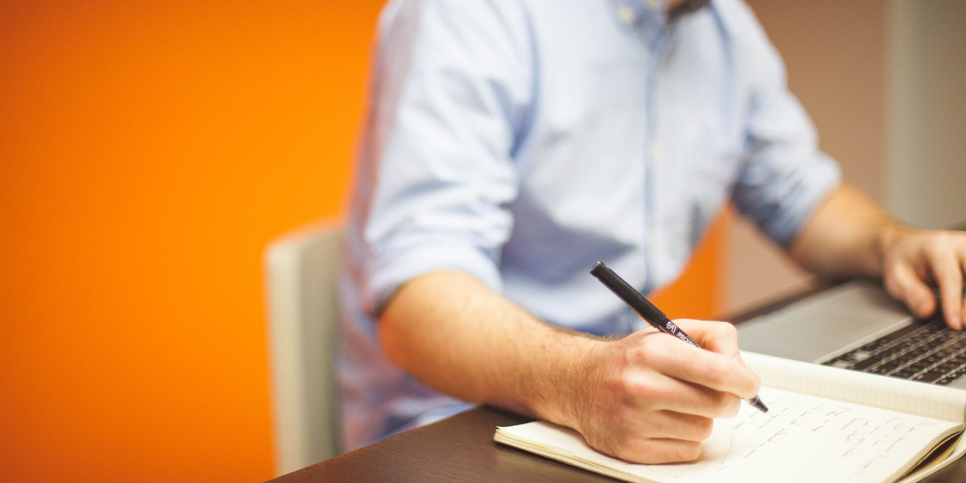 Person in a blue shirt writing in a notebook beside a laptop on a desk, orange wall behind.