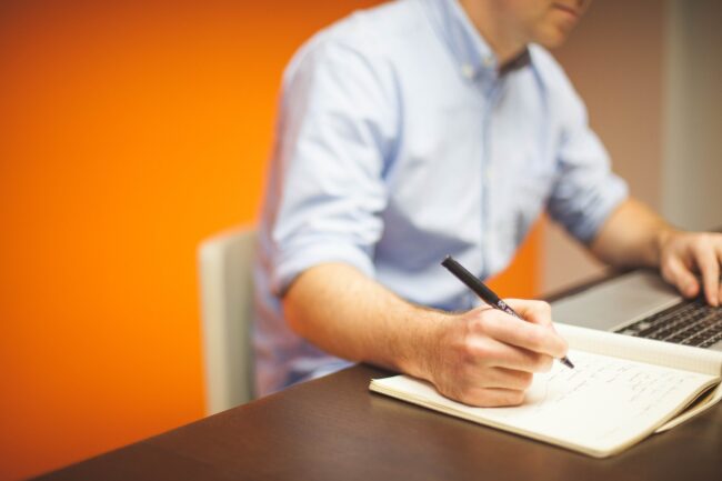 Person in a blue shirt writing in a notebook beside a laptop on a desk, orange wall behind.